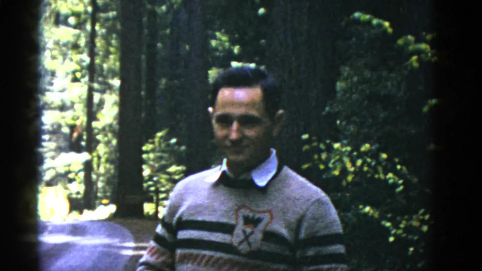 A man wearing a collared shirt and striped sweater stands in dappled forest light in this 1950s home-movie frame.