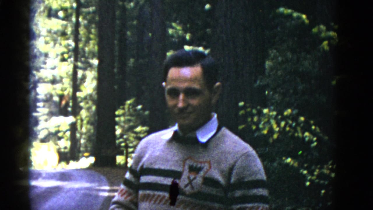 Man in a striped sweater smiles in a wooded setting beside a road.