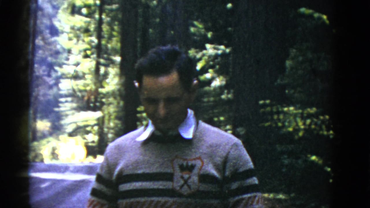 The man lowers his gaze in the wooded setting, with tall trunks, foliage, and a road visible behind him.