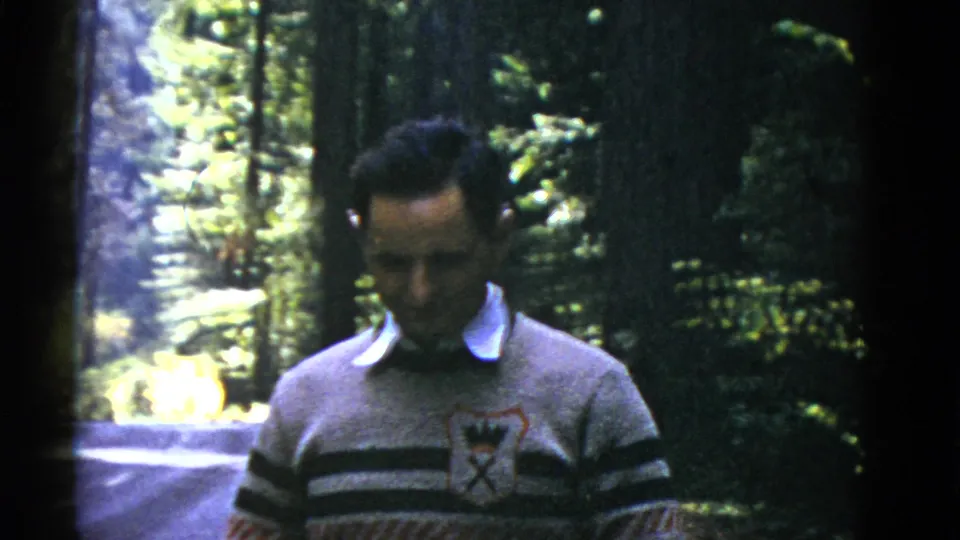 The man lowers his gaze in the wooded setting, with tall trunks, foliage, and a road visible behind him.