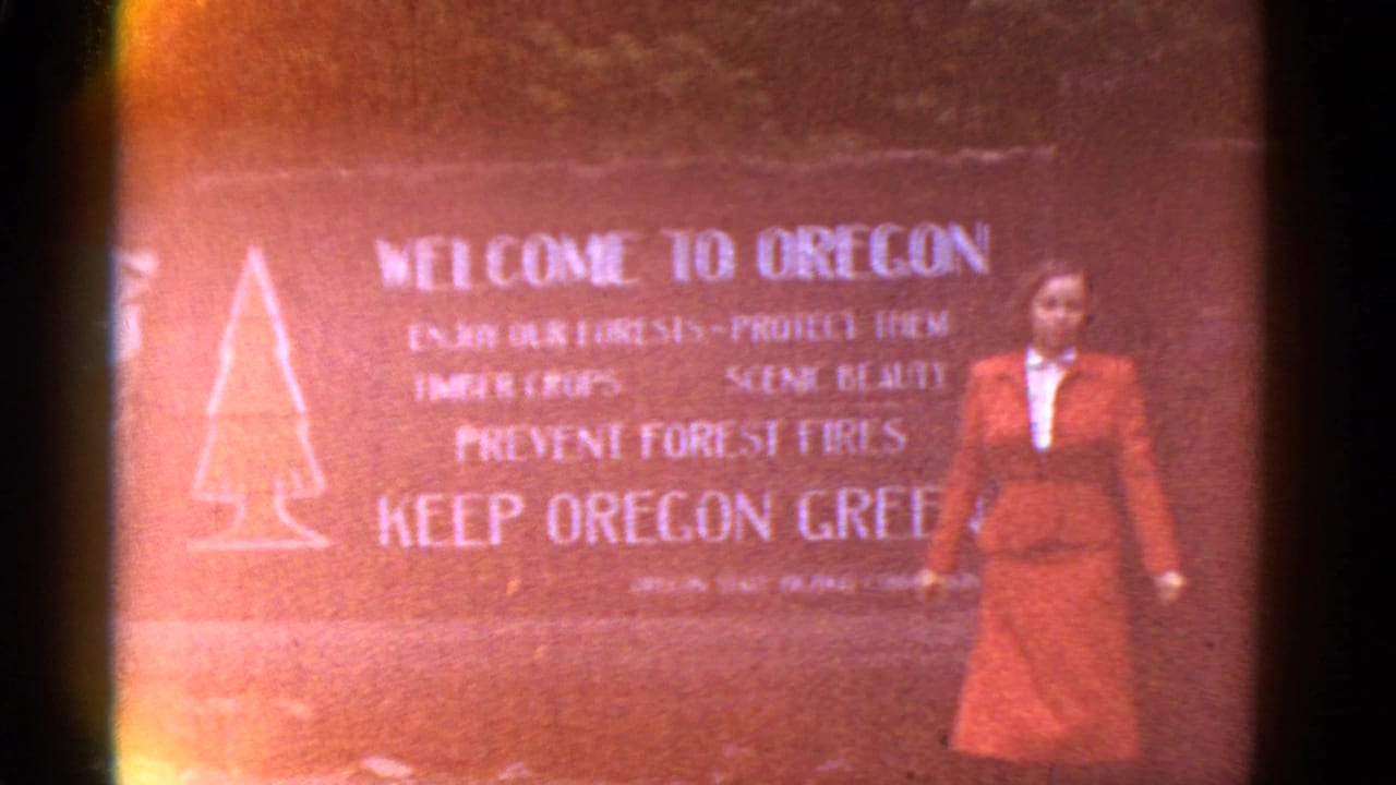 Woman in a red suit standing beside a Welcome to Oregon sign.