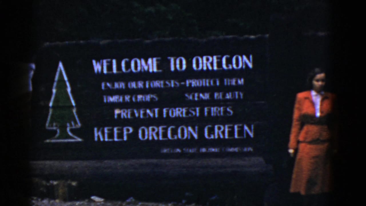 1950s 8mm frame of a woman standing near a Welcome to Oregon sign with forest-fire prevention text.