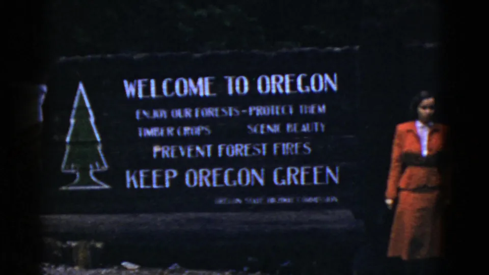 1950s 8mm frame of a woman standing near a Welcome to Oregon sign with forest-fire prevention text.