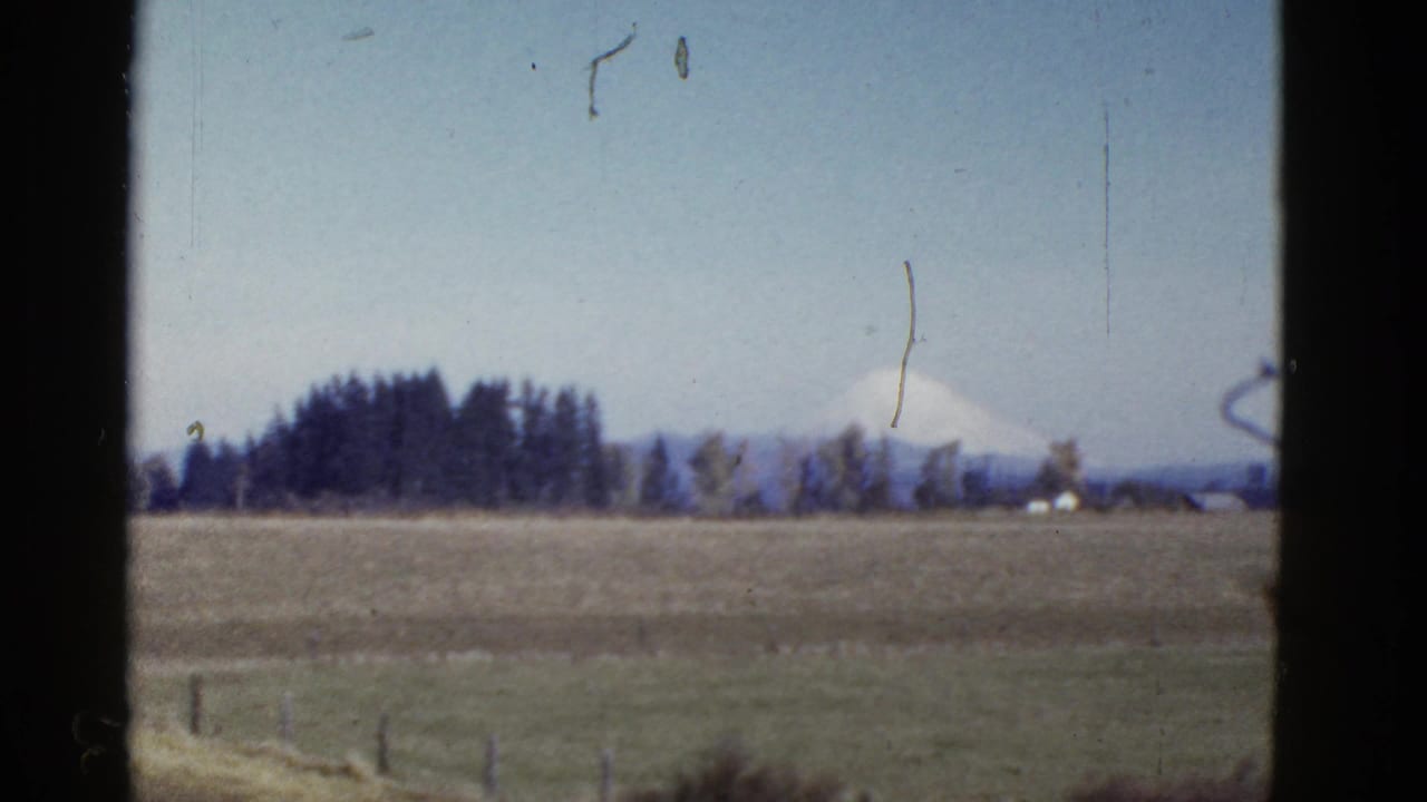 1950s 8mm frame showing a distant snow-capped mountain past open fields, fence posts, and a tree line.