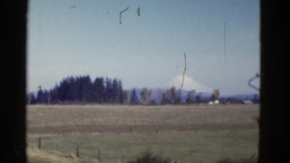 1950s 8mm frame showing a distant snow-capped mountain past open fields, fence posts, and a tree line.