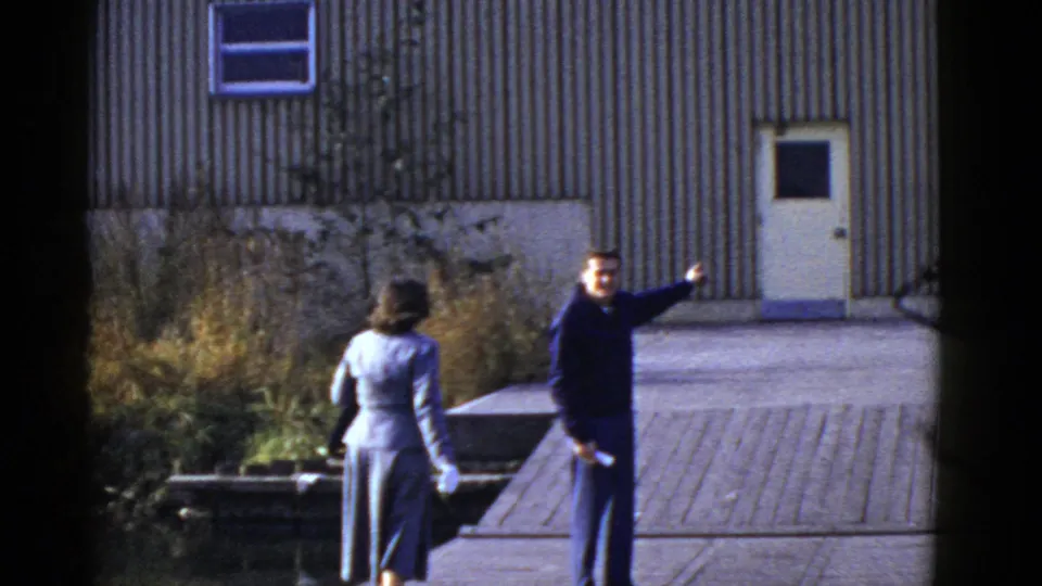 Two adults outside a corrugated metal building in 1950s home-movie footage, one gesturing toward the distance.