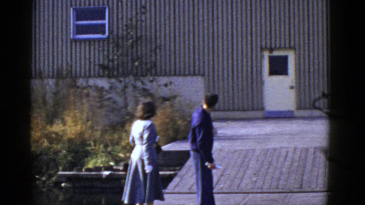 In San Francisco home-movie footage, a woman in a light suit and a man in dark clothing walk away near a corrugated metal wall.