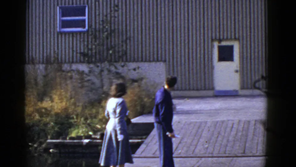 In San Francisco home-movie footage, a woman in a light suit and a man in dark clothing walk away near a corrugated metal wall.