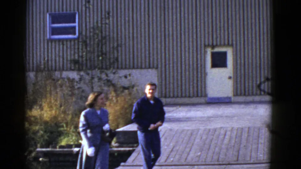 The couple turns toward the camera in the 1950s San Francisco home-movie scene, with the corrugated building still behind them.