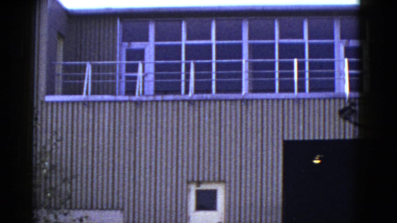 1950s San Francisco 8mm frame showing a utilitarian corrugated building facade with upper windows, railings, and lower doors.