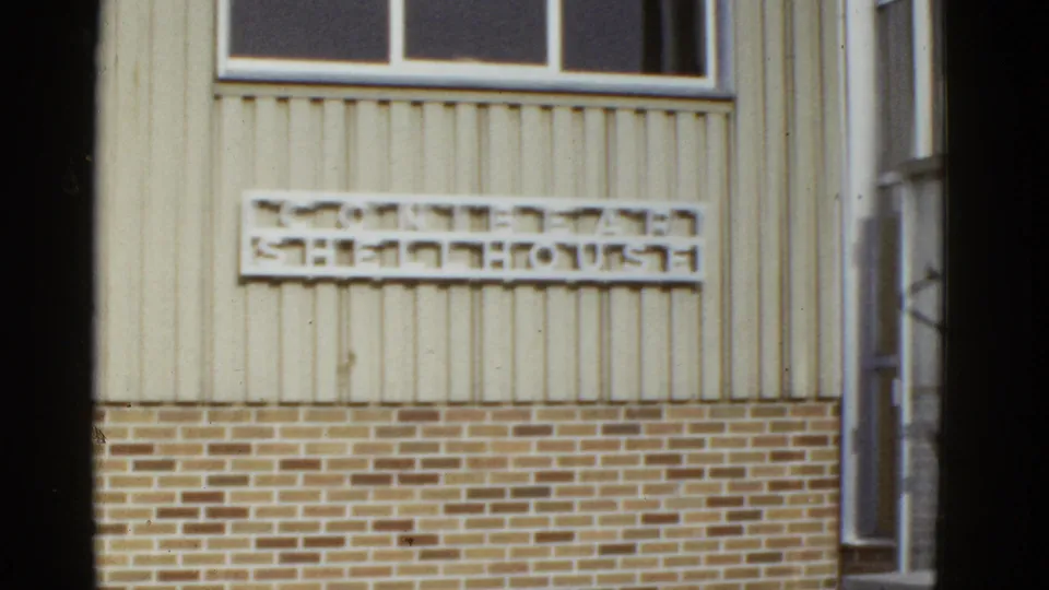 1950s San Francisco exterior showing light vertical siding, a brick lower wall, a window, and a blurred sign on the facade.