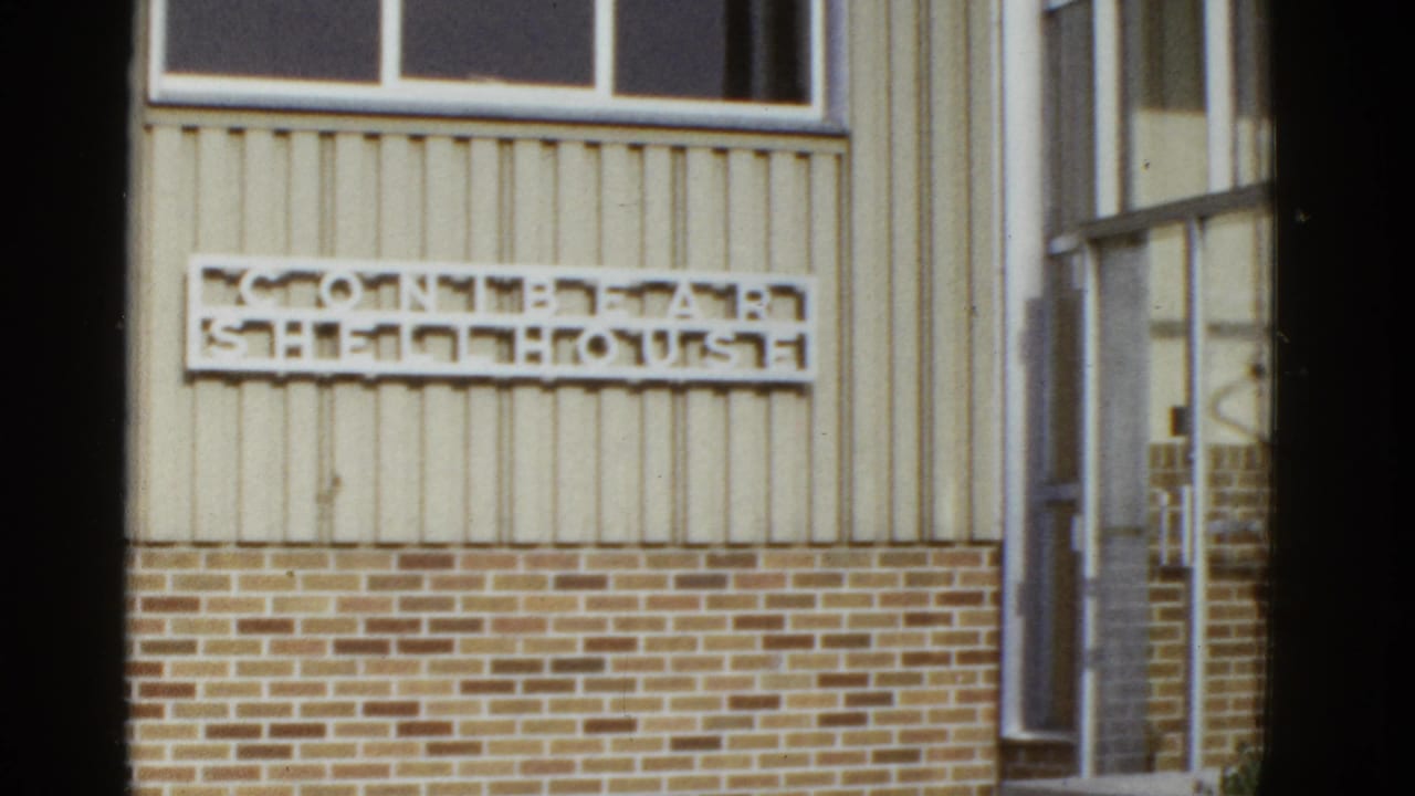 1950s San Francisco home-movie frame showing a mounted wall sign on beige siding above a brick foundation.