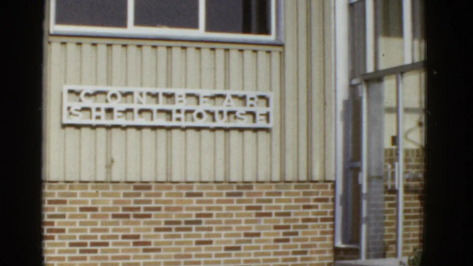 1950s San Francisco home-movie frame showing a mounted wall sign on beige siding above a brick foundation.