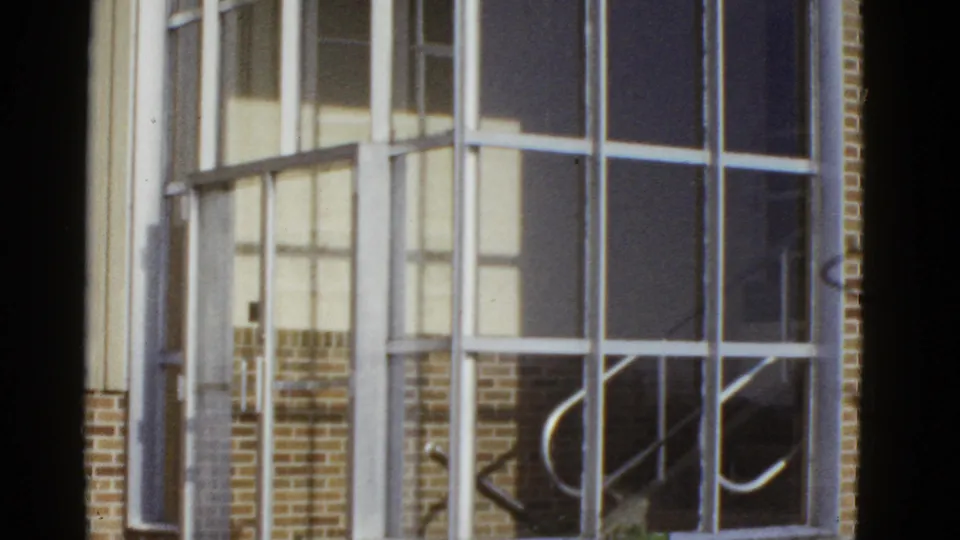 1950s San Francisco home-movie frame looking toward large window grids, brick facade, and reflected interior or stair details.