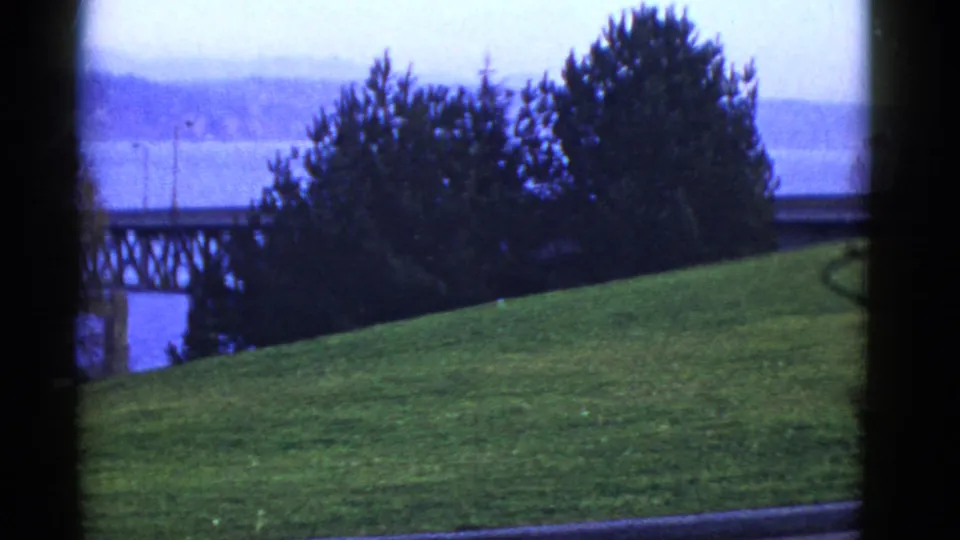 1950s San Francisco frame looking past a grassy hill and trees toward water, a distant bridge, and hazy hills beyond.