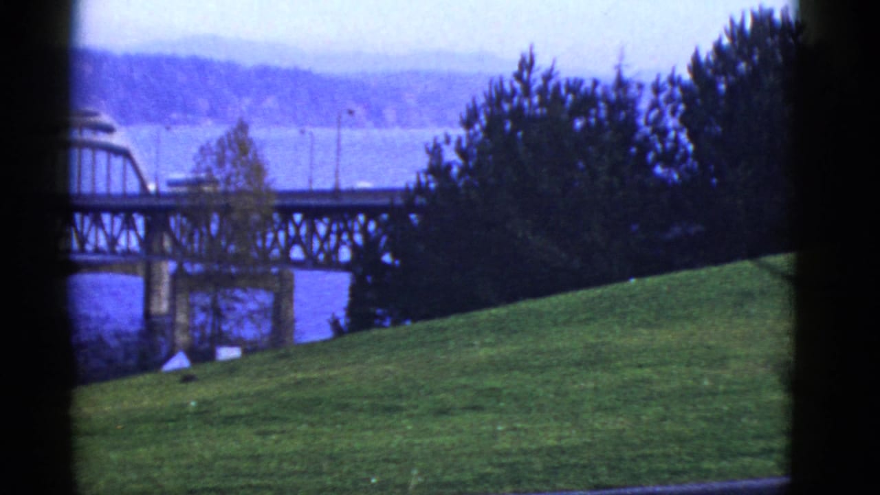 Early 1950s San Francisco-area view of a bridge crossing water, framed by dark edges with trees and a grassy slope in front.