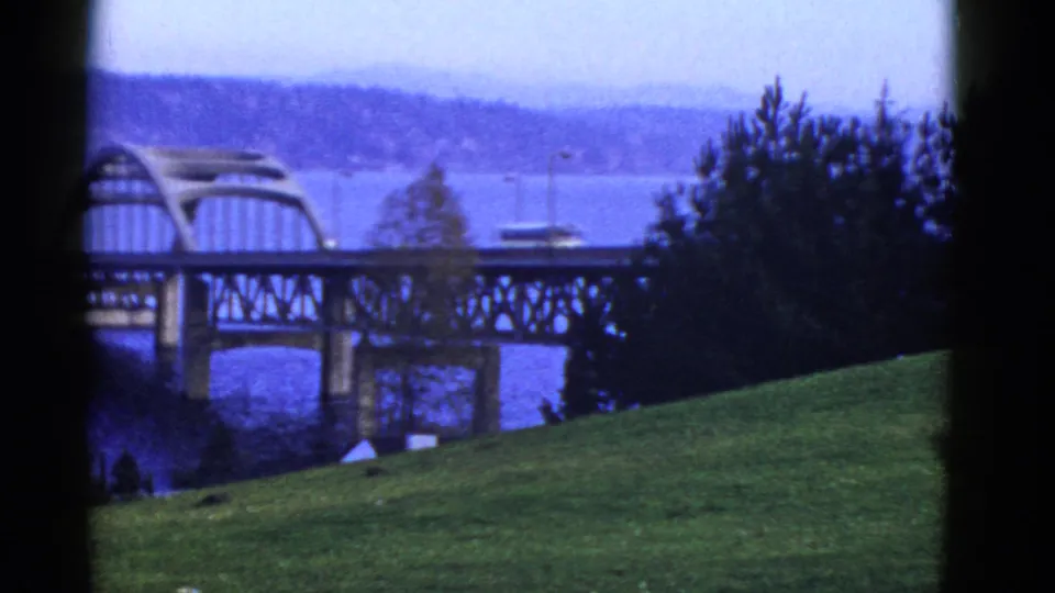 Early 1950s San Francisco-area bridge view with an arched structure, water below, green slope, trees, and a hazy distant shoreline.
