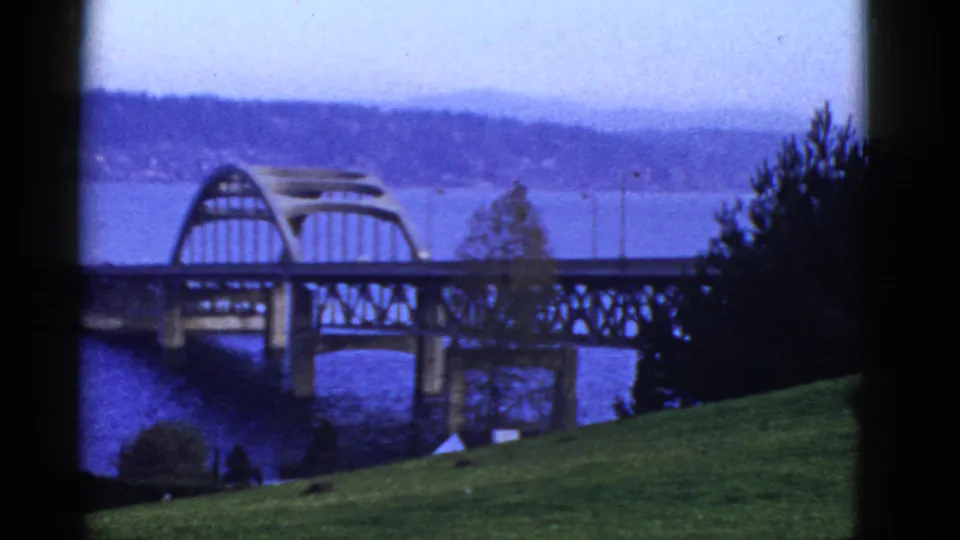Early 1950s San Francisco-area 8mm frame showing the arched bridge, water, grassy foreground, trees, and distant hills under haze.