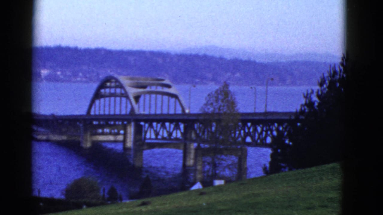 1950s 8mm home-movie view of an arched bridge over blue bay water near San Francisco, with distant hills and dark frame edges.