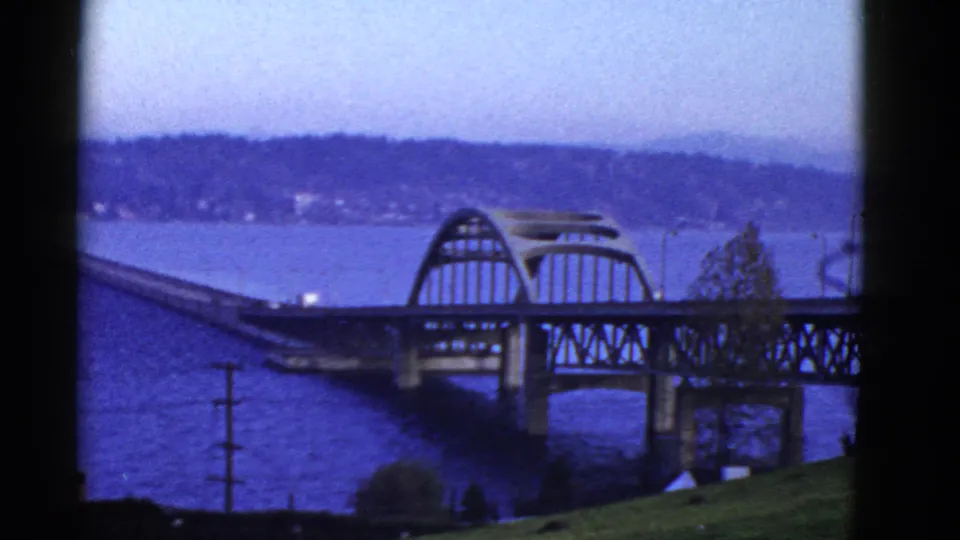 The bridge approach leads toward an arched span across blue water, with a distant landmass and soft 8mm color shift.