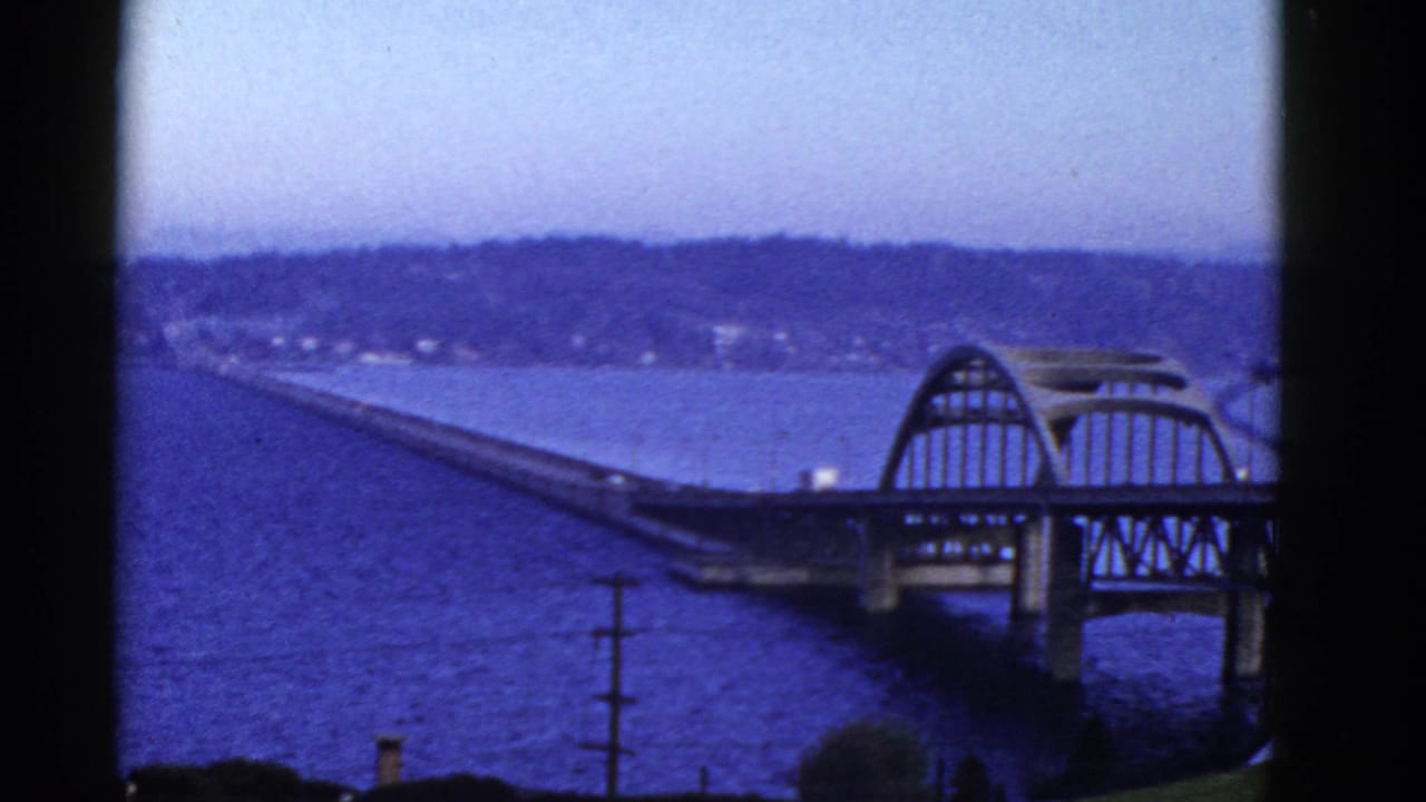 1950s 8mm view of a San Francisco-area bridge over blue water, with an arched section, shoreline hillside, and dark frame borders.