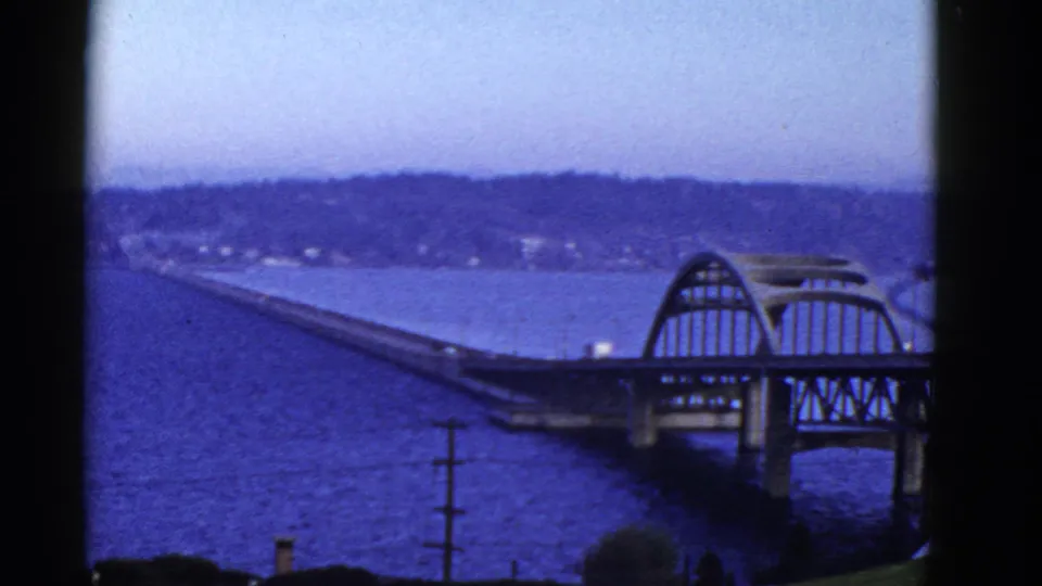 1950s 8mm view of a San Francisco-area bridge over blue water, with an arched section, shoreline hillside, and dark frame borders.