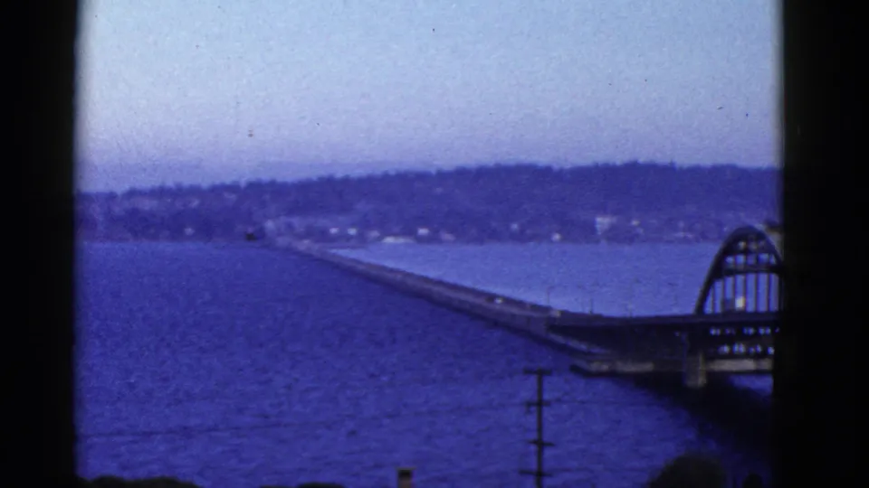 Elevated 1950s home-movie view of a long San Francisco-area bridge crossing blue water toward a tree-lined shore.