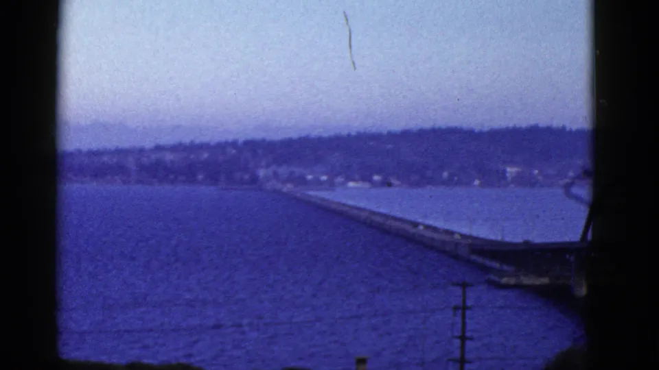 San Francisco-area water crossing seen from above, with a long bridge approach, distant hillside, and blue-tinted 8mm film texture.