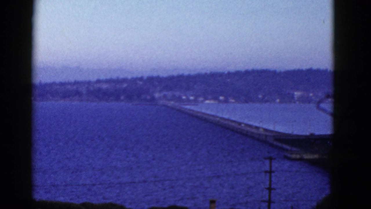 1950s 8mm frame showing a blue-toned bay, distant hills, and a long bridge or pier-like structure seen through dark film borders.