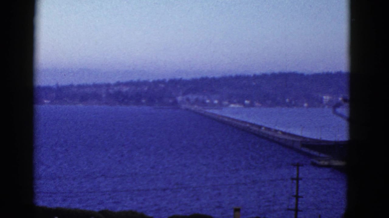 Blue-toned 8mm view of a bay and distant shoreline, with a long bridge or pier-like span crossing part of the water.