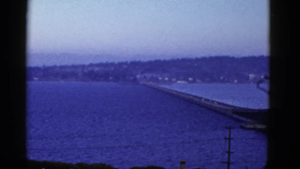 Blue-toned 8mm view of a bay and distant shoreline, with a long bridge or pier-like span crossing part of the water.