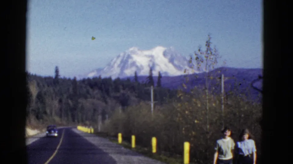 1950s home-movie road scene with a period car, roadside posts, trees, two pedestrians, and a snow-capped mountain in the distance.