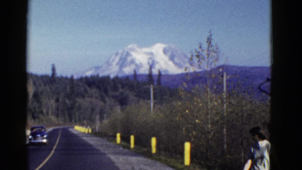 Early 1950s home-movie frame of a paved road through trees toward a snow-capped mountain, with a car and roadside pedestrian visible.