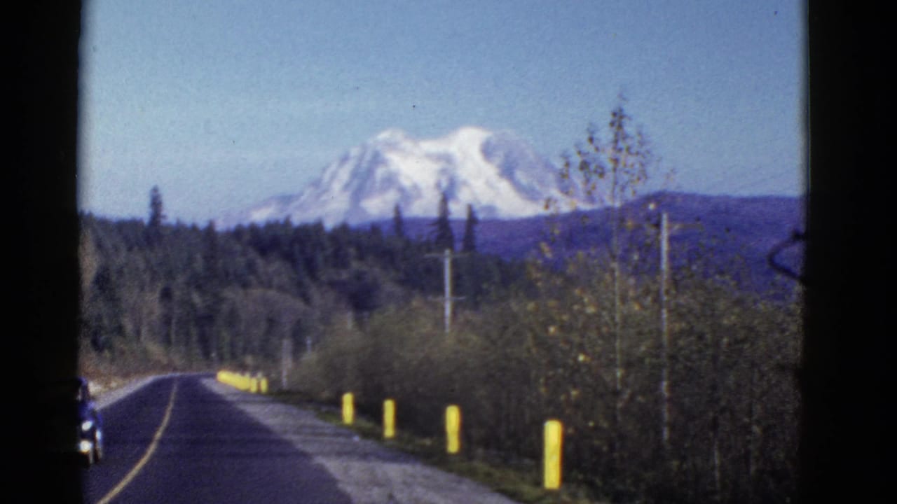 Early 1950s 8mm frame of a forest-lined road leading toward a distant snow-capped mountain under a blue sky.