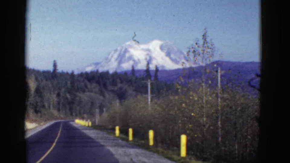 Early 1950s road-trip frame showing a paved road and yellow markers below a distant snow-capped mountain, with trees and utility poles nearby.
