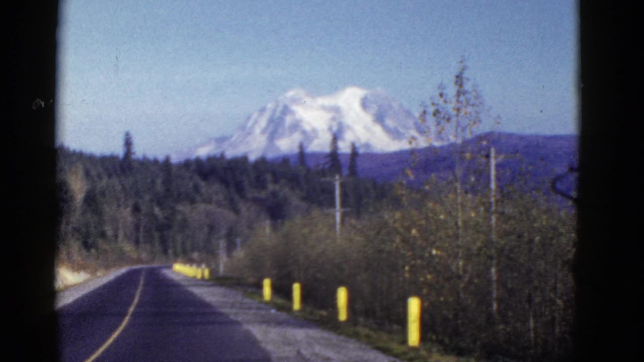 1950s 8mm travel frame showing a paved road, forest, utility poles, and a distant snow-capped mountain near Mount Rainier.