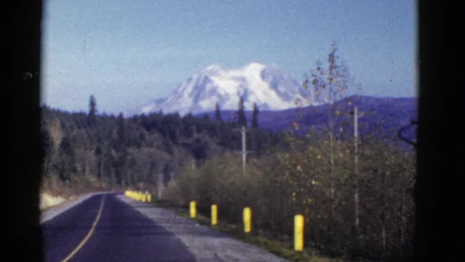 1950s 8mm travel frame showing a paved road, forest, utility poles, and a distant snow-capped mountain near Mount Rainier.