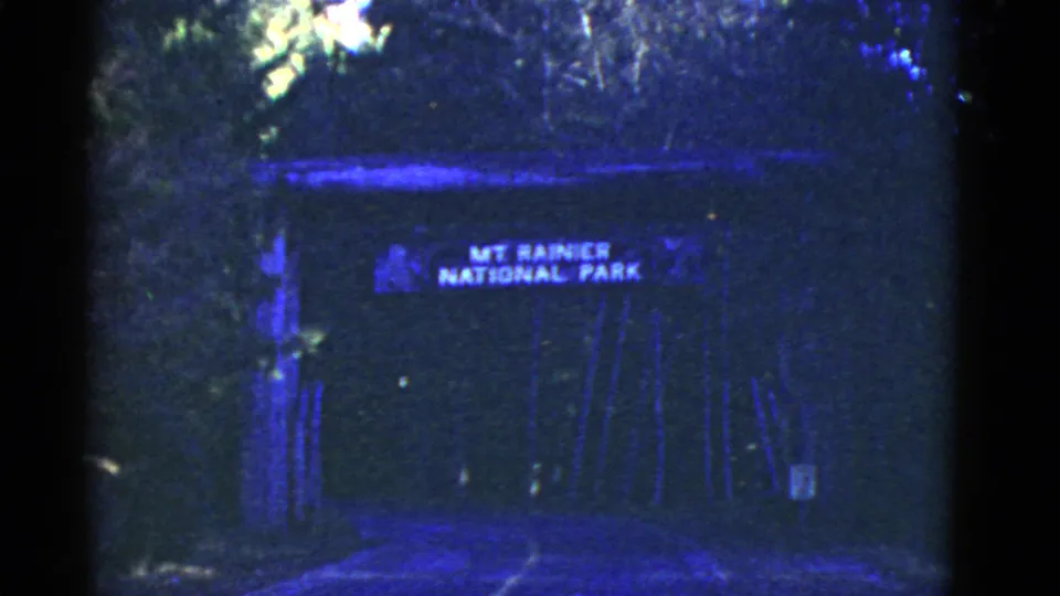 1950s 8mm frame with the Mount Rainier National Park sign over a dark, tree-lined gateway or tunnel approach.