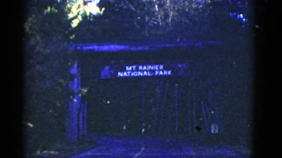 Later frame of the Mount Rainier National Park gateway, with heavy blue cast, dark road surface, and surrounding trees.