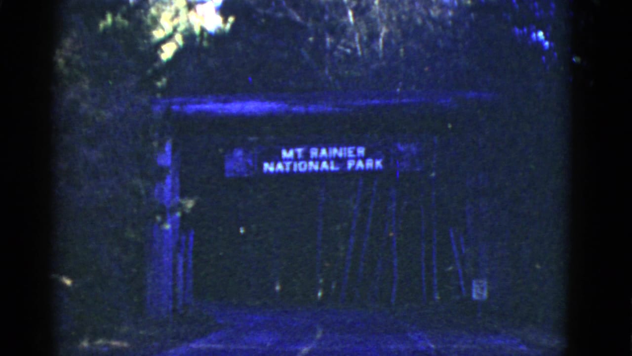 A dark 8mm frame shows a Mount Rainier National Park sign set back among trees, with heavy shadow and blue color cast.