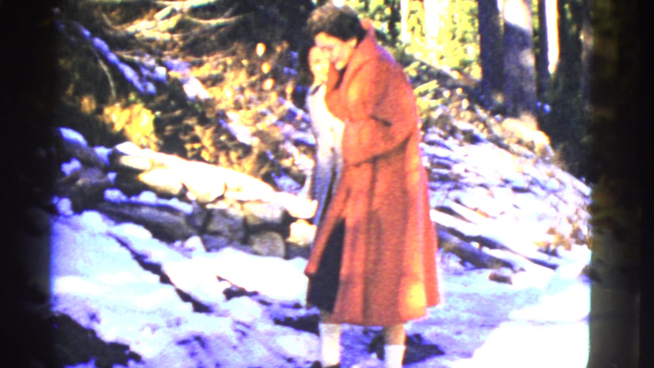 A woman in a long red coat carefully stands on snowy ground in the Mount Rainier park area, with rocks and forest behind her.