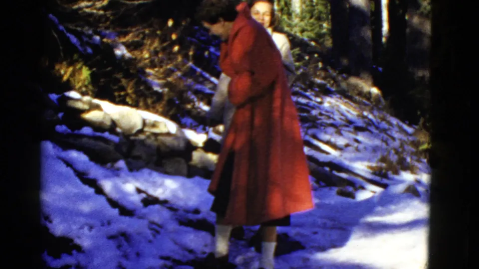 Two women appear on a snowy park path, one in a red coat stepping carefully while another stands partly behind her.