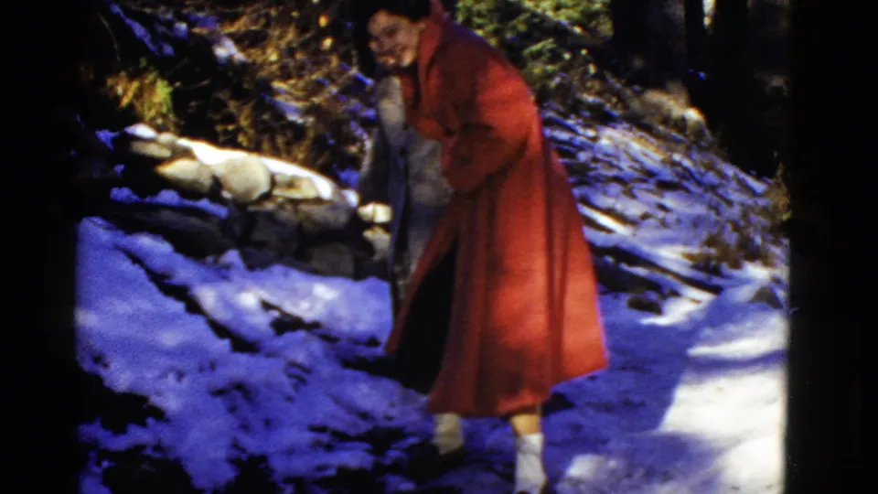 A woman in a long red coat bends or steps cautiously on snow, with another woman partly visible behind her among rocks and trees.