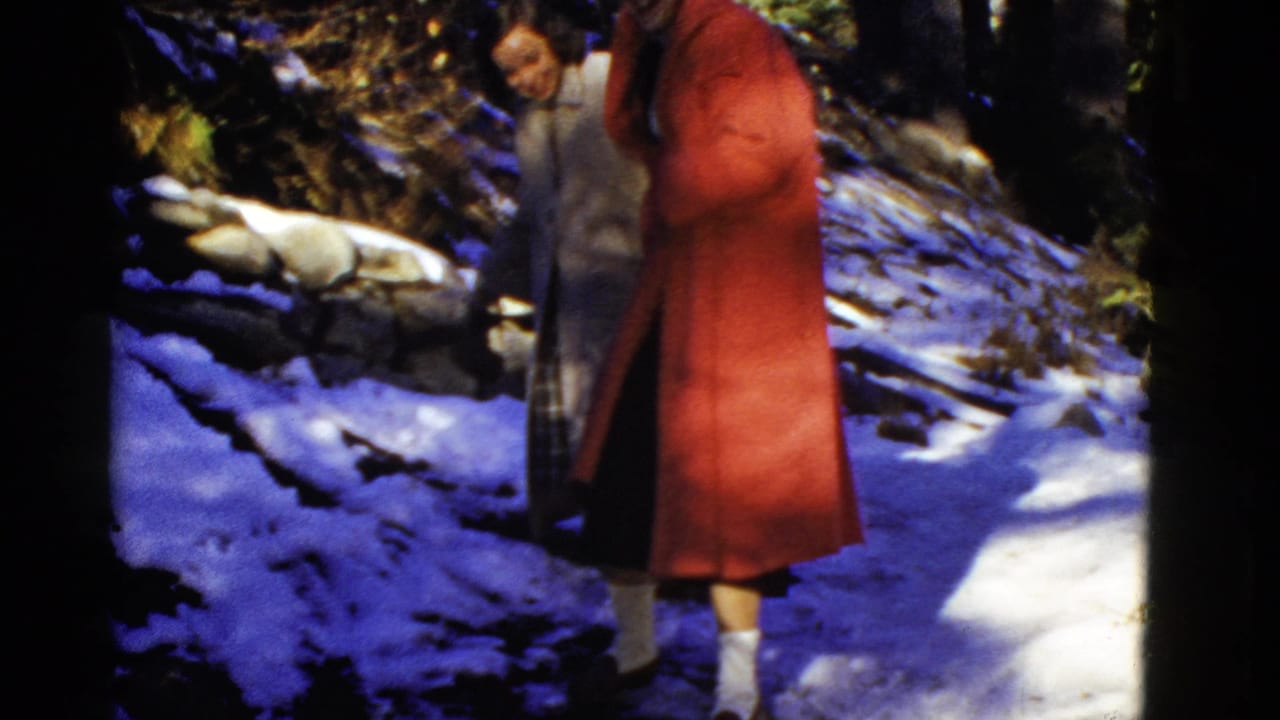 Early 1950s 8mm home-movie frame of two adults on a snow-covered wooded path, with a red coat prominent in the foreground.