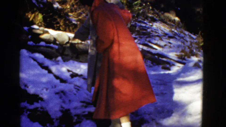 Early 1950s 8mm home-movie frame of a red-coated adult walking away along a snow-covered path in wooded shade.