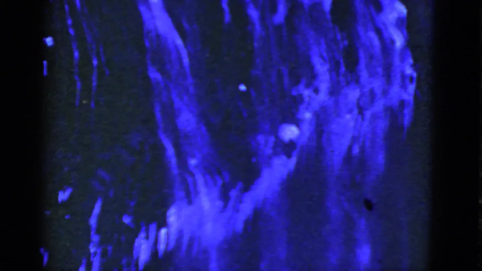 Abstract 8mm frame with blue vertical forms, a brighter lower patch, and dark surrounding space.