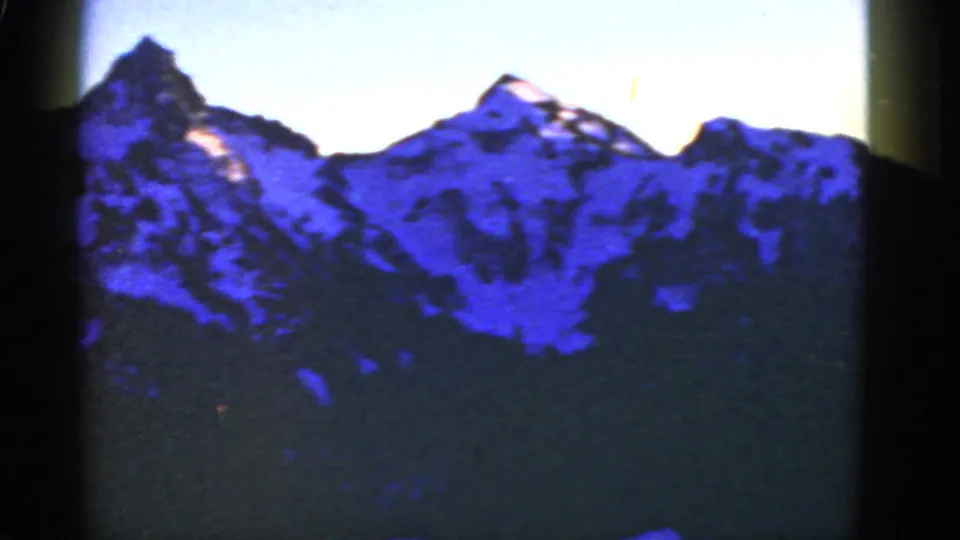 Scenic 8mm frame of rugged snowy mountain peaks above dark trees, with visible gate edge and blue color cast.