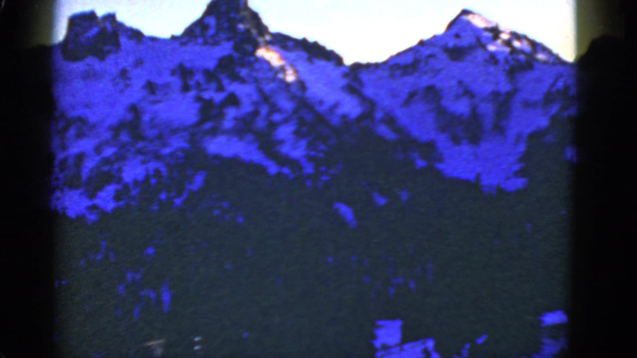 1950s 8mm frame of snowy mountain ridges and evergreen forest with strong blue color shift and visible film-edge darkness.