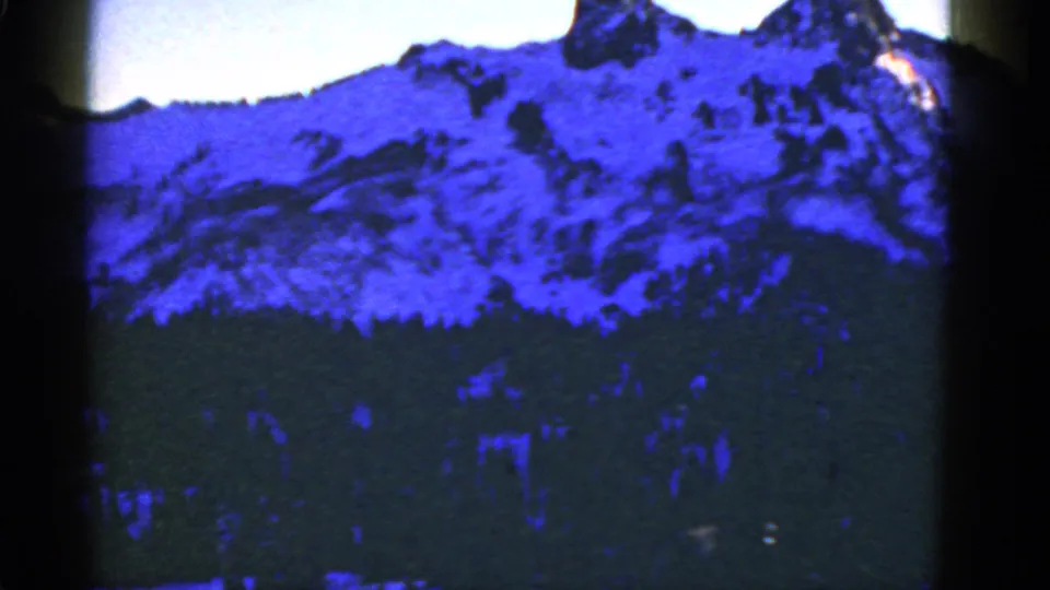 Archival 8mm frame showing a blue-toned snowy mountain slope with a dark forest band below and soft home-movie texture.