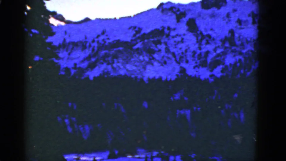 1950s 8mm landscape frame of a blue-shifted snowy mountain wall with forest below and visible film gate shadows.